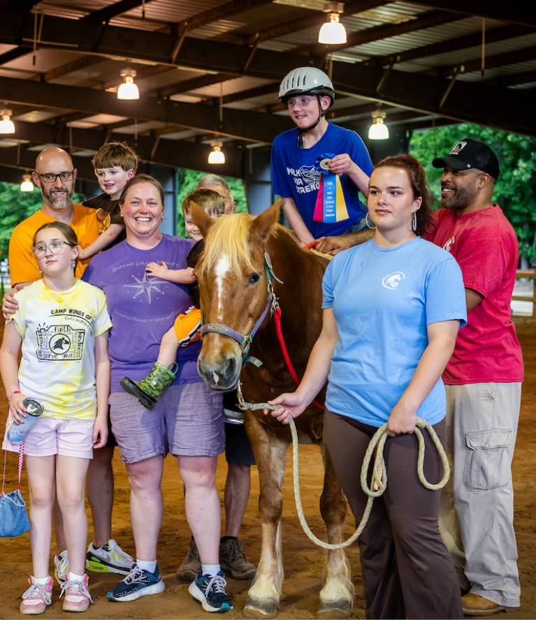 Group of children and volunteers gathered around a therapy horse during a therapeutic riding horse show inside an indoor arena.